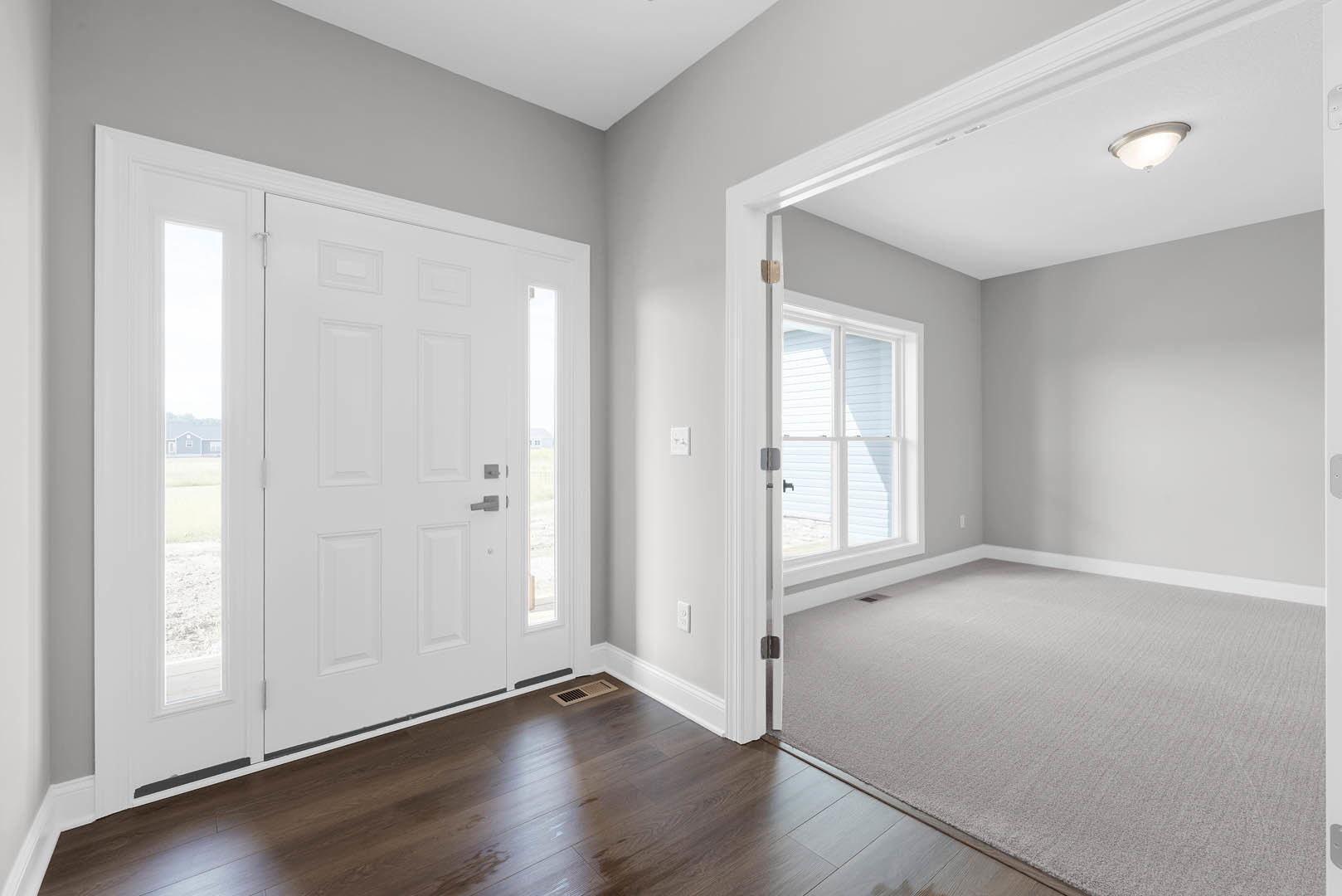 Carpeted room with white paneled doors, white trim, and a ceiling light fixture; window with white framing and wood floor vent visible.