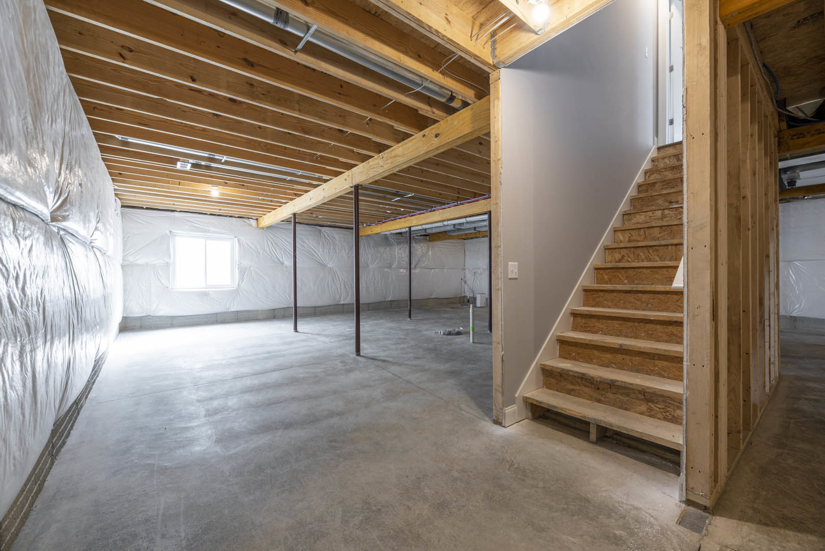 Open-concept room featuring wooden staircase with light wood steps, white plaster walls, exposed wooden ceiling beams, and a large window letting in bright natural light