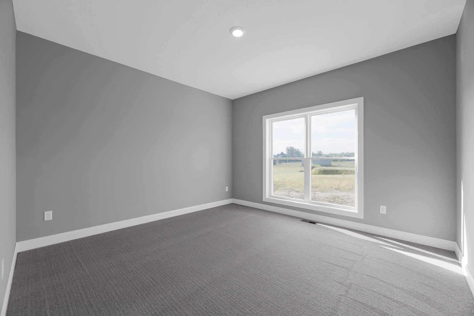 Bright room with white walls, large window overlooking grassy field, grey carpet flooring, recessed ceiling light, simple baseboard molding