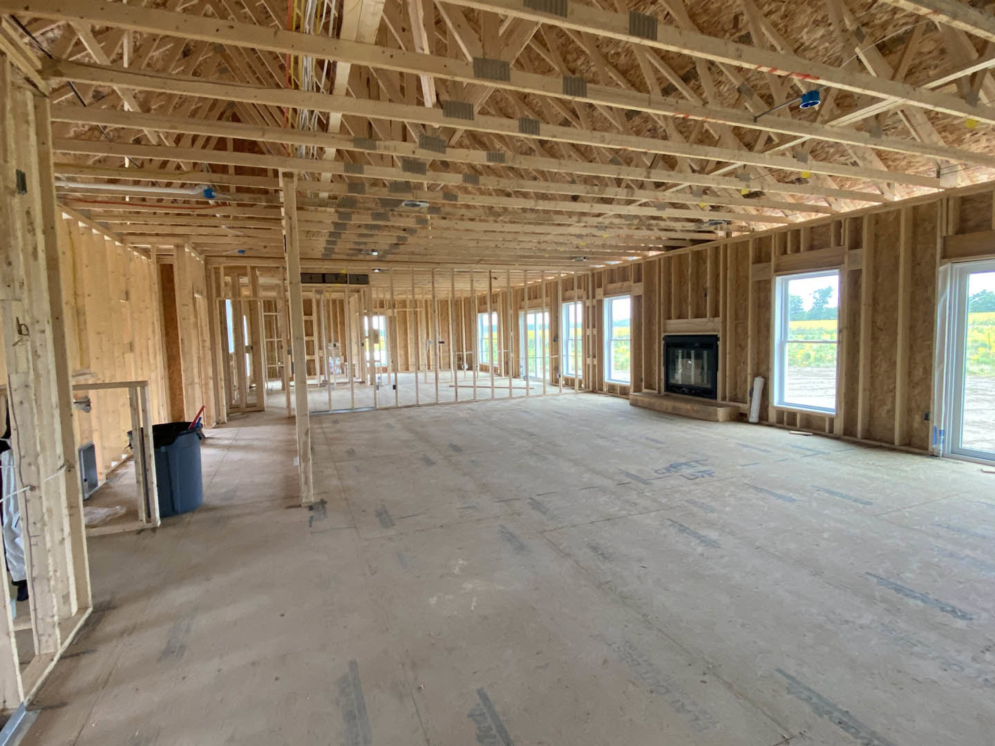 Living room with exposed wooden ceiling beams, concrete floor, large windows overlooking a field of trees, and a modern fireplace.