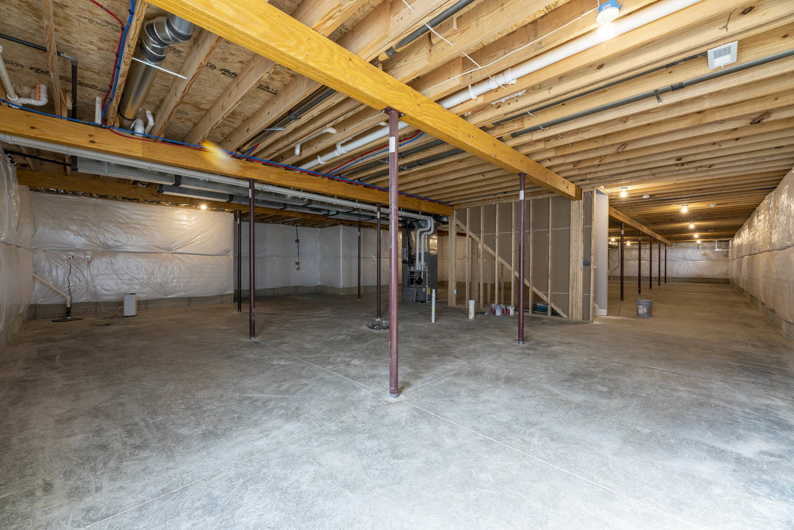 Exposed wooden ceiling beams and metal pipes above a concrete floor in a basement or unfinished room, with a white vent on wood and visible building insulation.