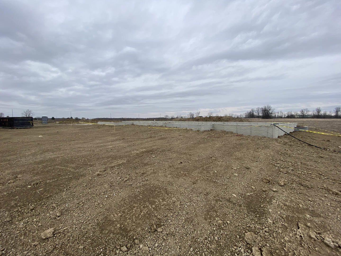 Large dirt field bordered by a concrete wall, leafless tree in the background, cloudy sky overhead, man standing on bare ground near the wall