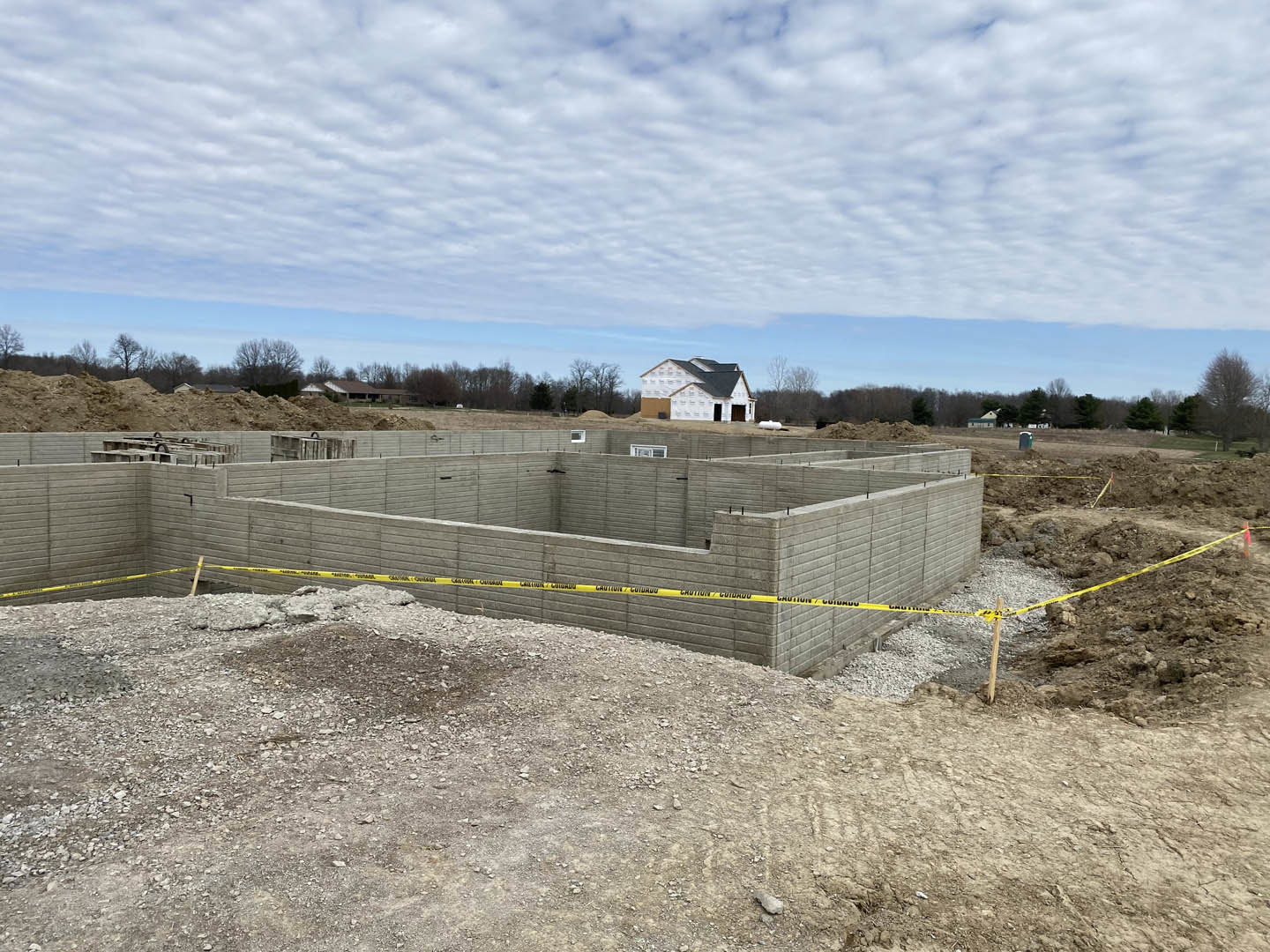 White house with black roof under construction, concrete wall with yellow tape in foreground, leafless tree, cloudy sky, grassy lot.