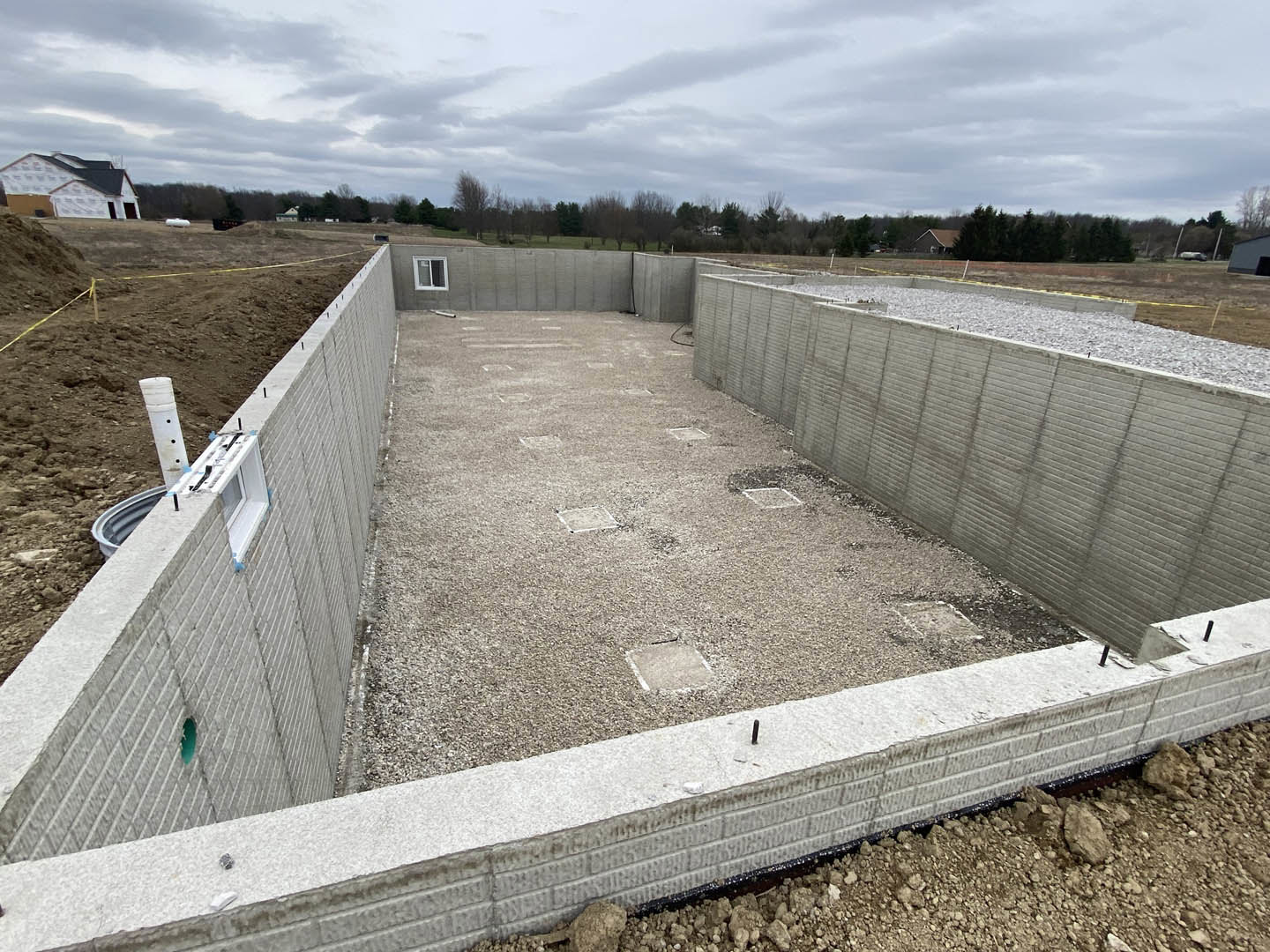 Concrete foundation with exposed hole, gravel base, and partial window opening, surrounded by soil and grass under cloudy sky with trees