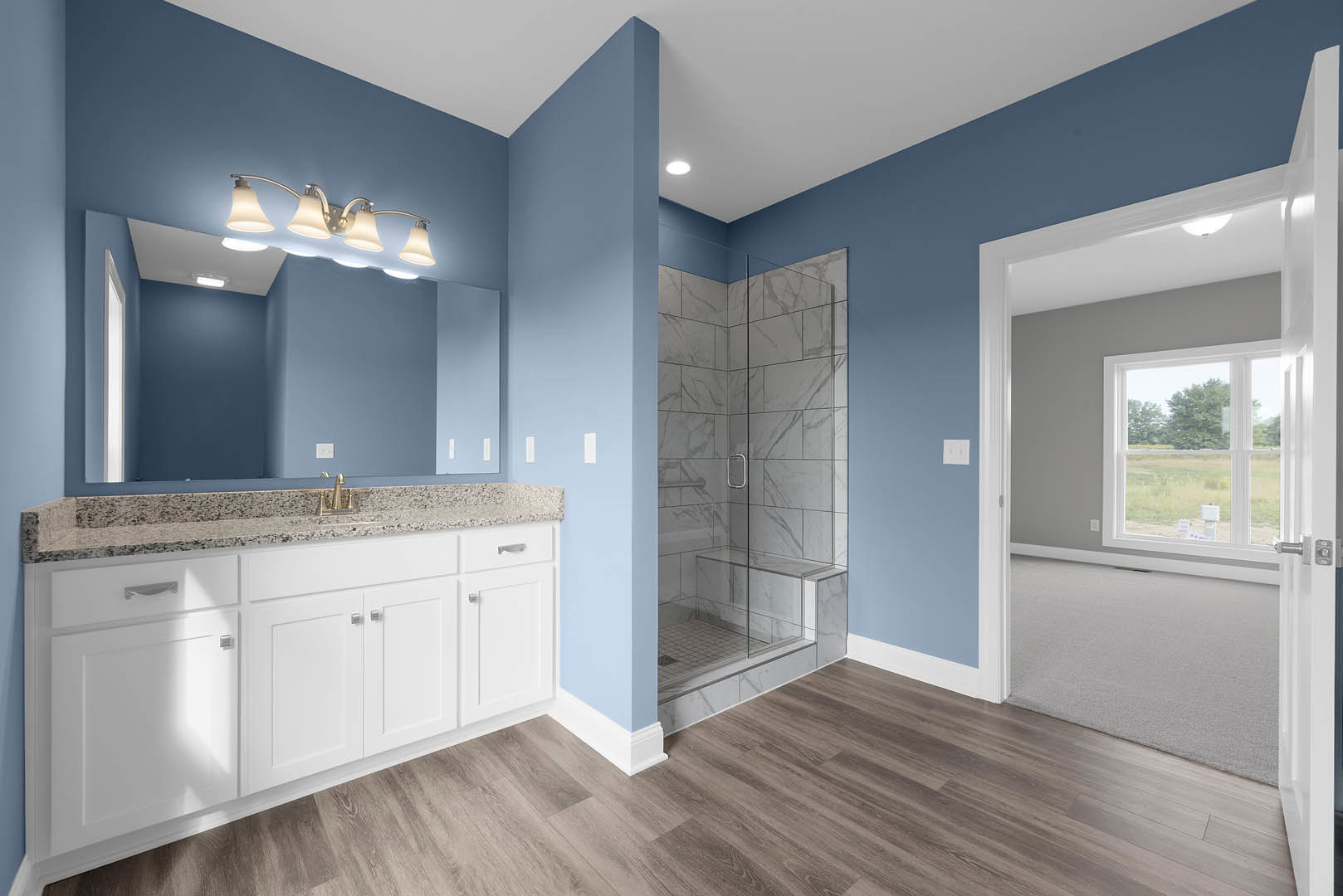 Bathroom with glass shower door, white tile shower wall, wood flooring, white baseboards, wall-mounted mirror, multi-bulb light fixture, and window overlooking a grassy field