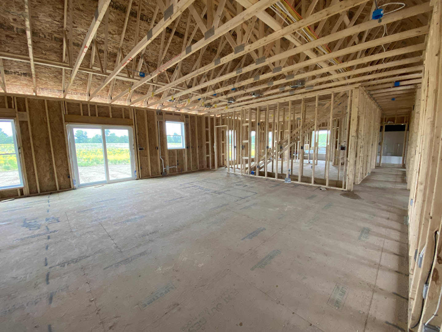 Open-concept room with exposed wooden ceiling beams, large window overlooking field and trees, light wood flooring, neutral walls