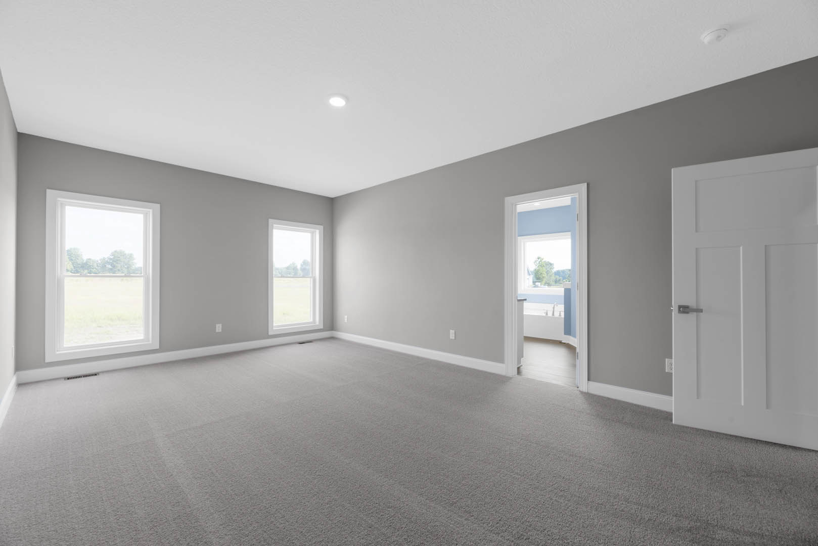 Grey-walled room with white trim, carpeted floor, white door with silver handle, large window overlooking grassy field