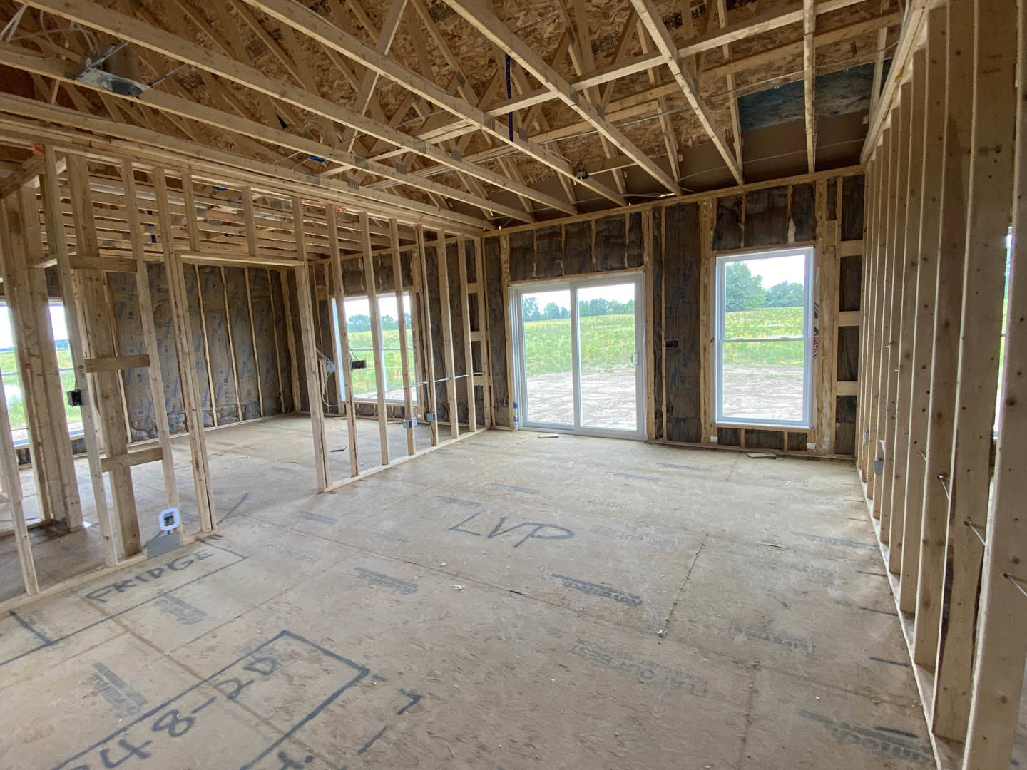 Wood-framed room under construction with exposed ceiling beams, multiple windows, and sunlight casting shadows on unfinished floor