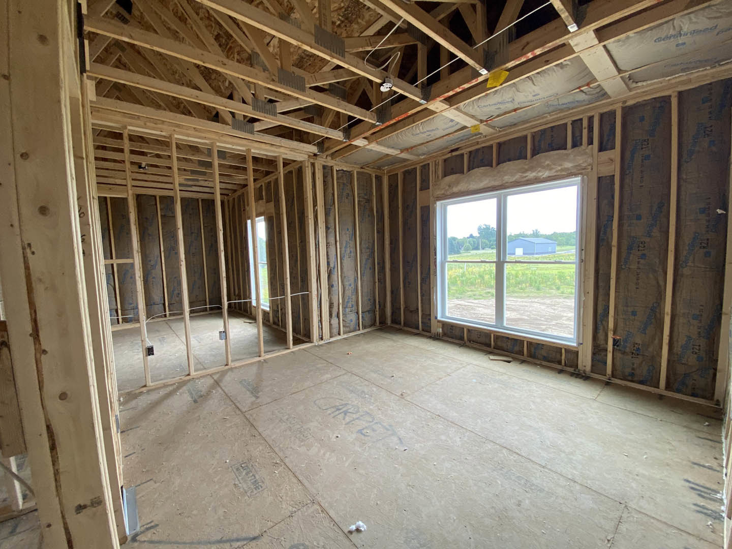 Wood-framed window overlooking blue barn, exposed wooden ceiling beams, concrete floor, and unfinished walls in custom home interior