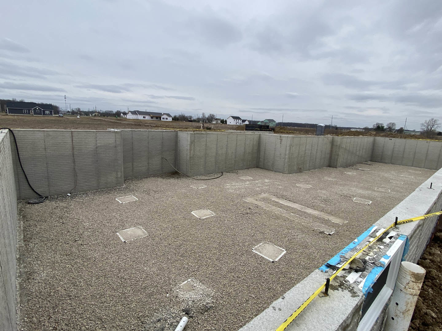 Concrete foundation with square blocks, white rectangular object on dirt ground, cloudy sky above open field, close-up of pipe near composite exterior wall
