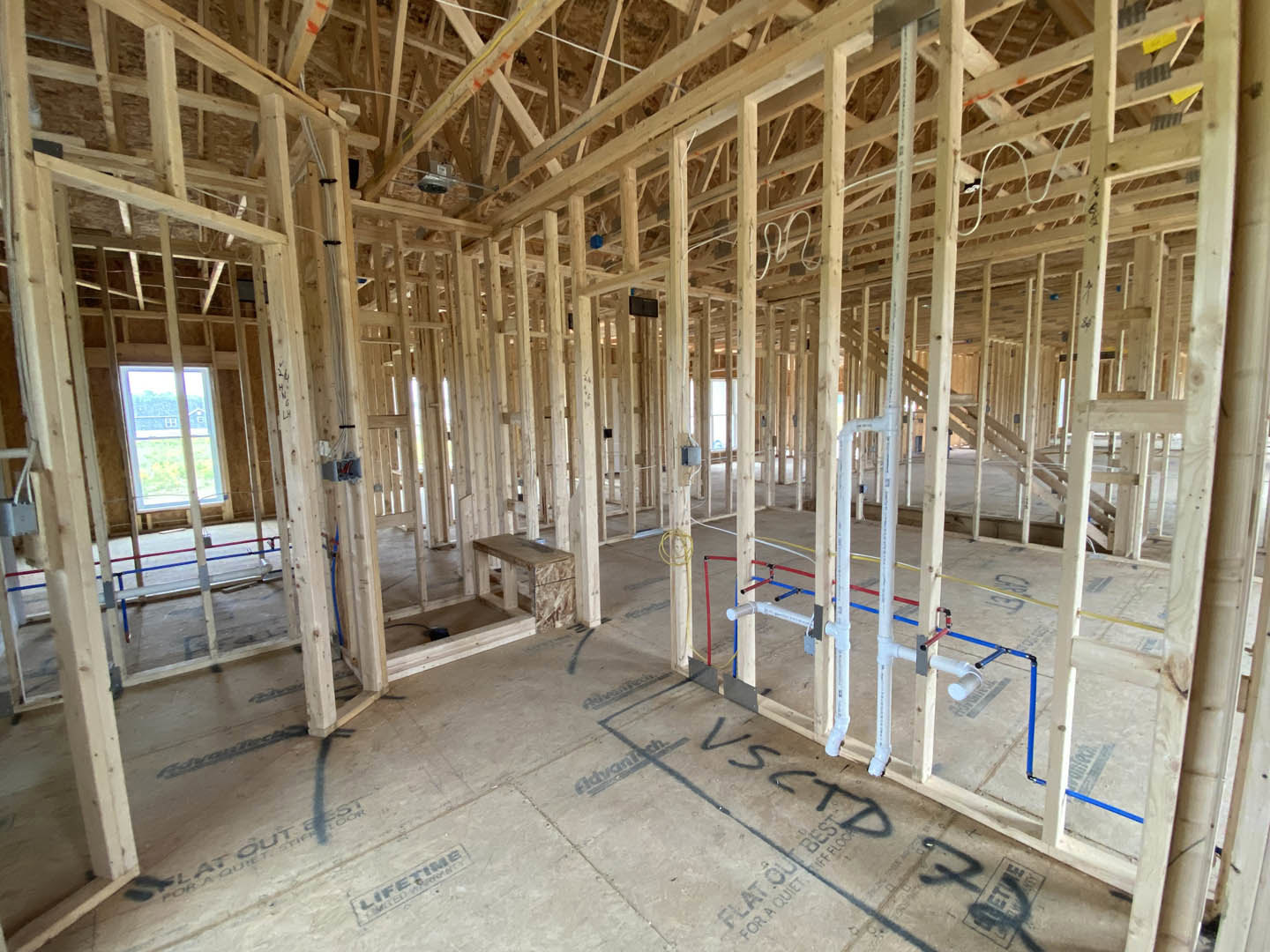 Exposed wood framing with vertical beams, unfinished plywood floor, visible plumbing pipes, and open window cutouts in a house under construction