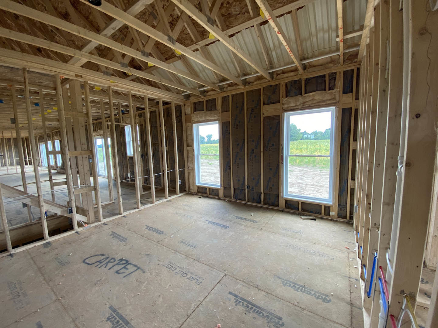 Living room with exposed wood ceiling beams, large windows overlooking a grassy field, unfinished wood framing, and natural daylight streaming onto the floor.