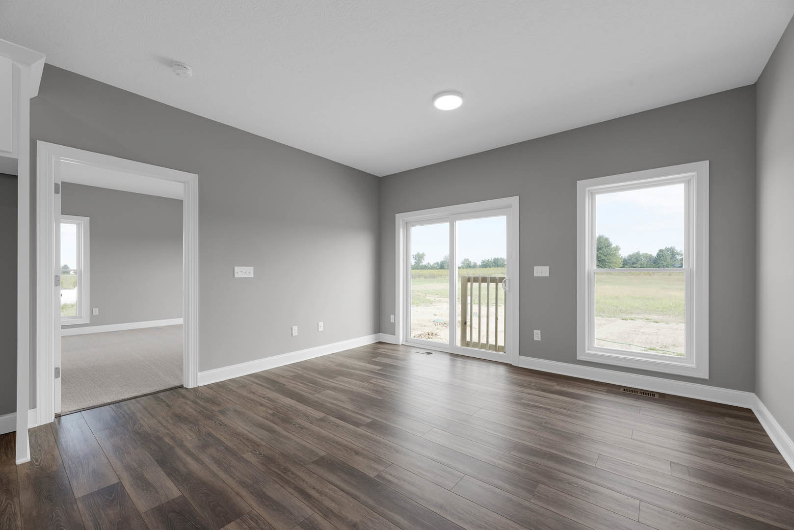 Open room with wide plank wood flooring, large windows and sliding glass door overlooking grassy field, ceiling light fixture, and open doorway.