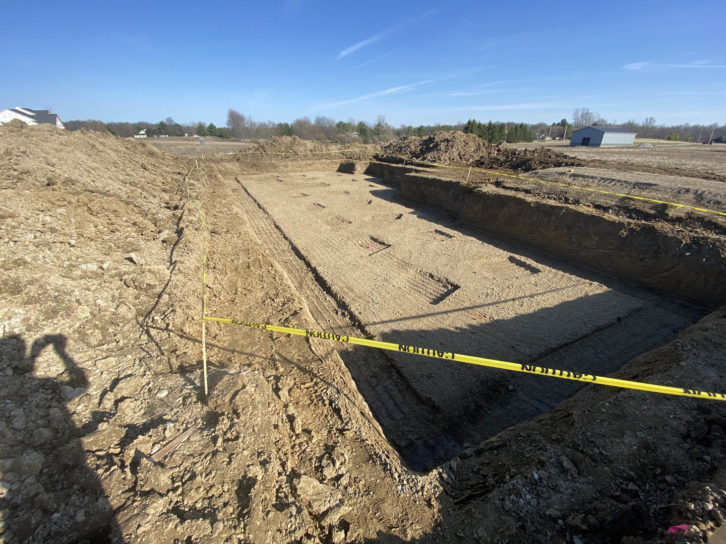 Yellow caution tape stretched across a residential construction site, partially built roof with dark shingles, exposed soil and foundation, blue sky with scattered white clouds
