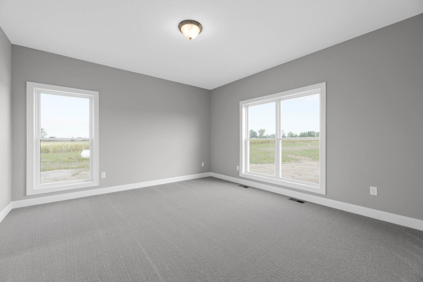 Bright room with large windows overlooking a grassy field, grey carpet flooring, white plaster walls, ceiling light fixture, and crown molding.