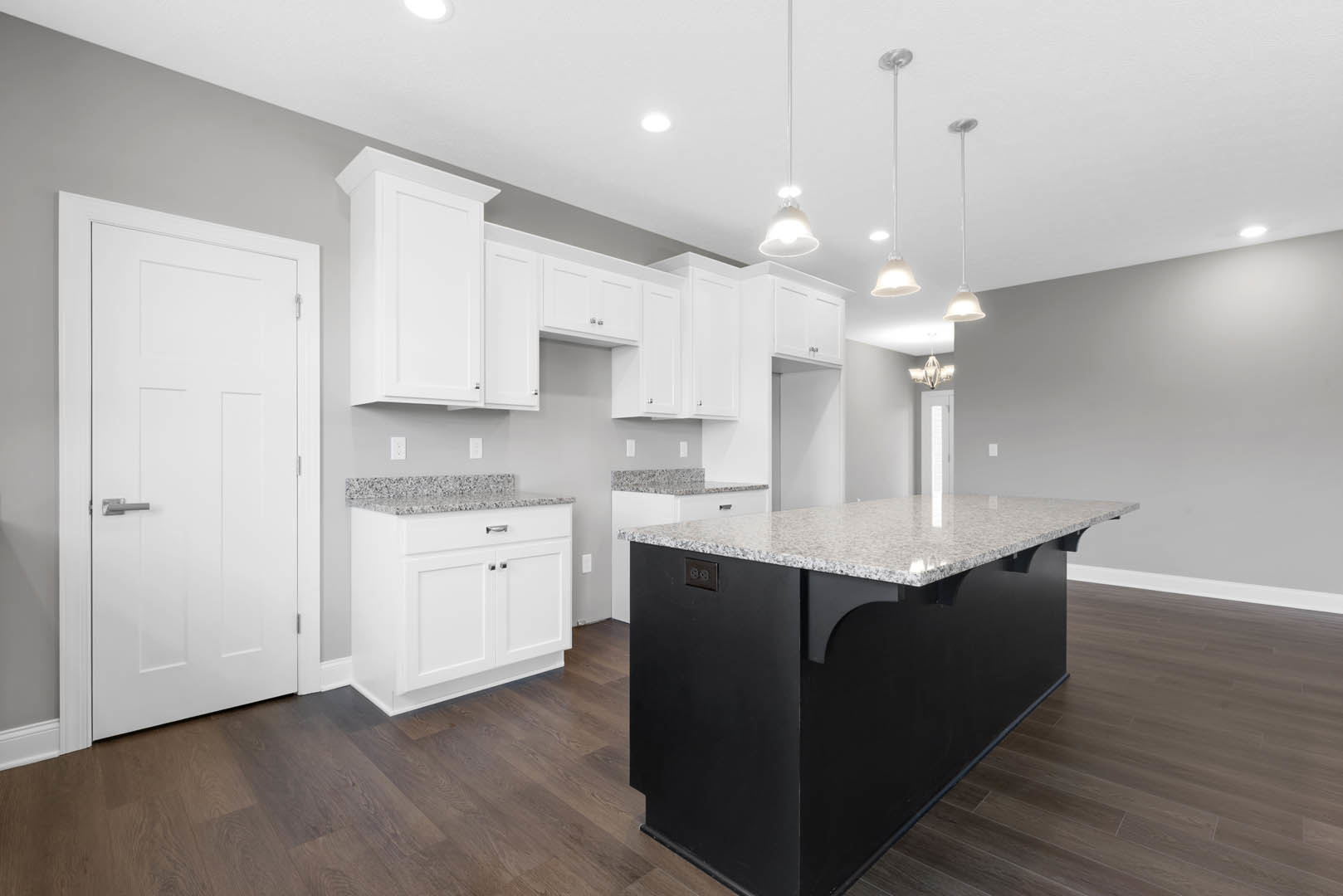 Black kitchen island with white cabinets, granite countertops, stainless steel sink, silver cabinet handles, pendant light fixture, hardwood flooring, and white door in background