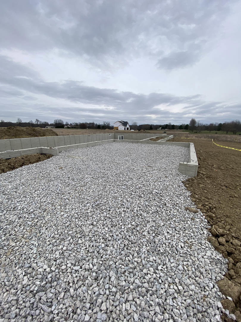 Gravel road bordered by grass and wildflowers, cloudy sky overhead, yellow caution tape stretched across dirt path, concrete retaining wall lined with rocks