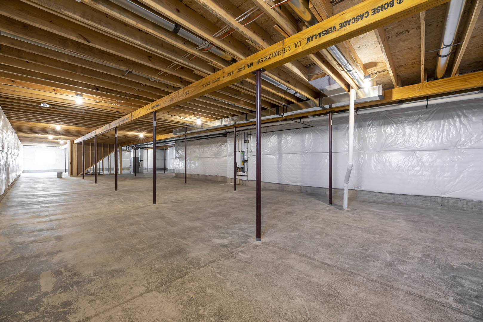 Spacious interior with exposed wooden ceiling beams, white wall partially covered in plastic, concrete floor, and steel support poles