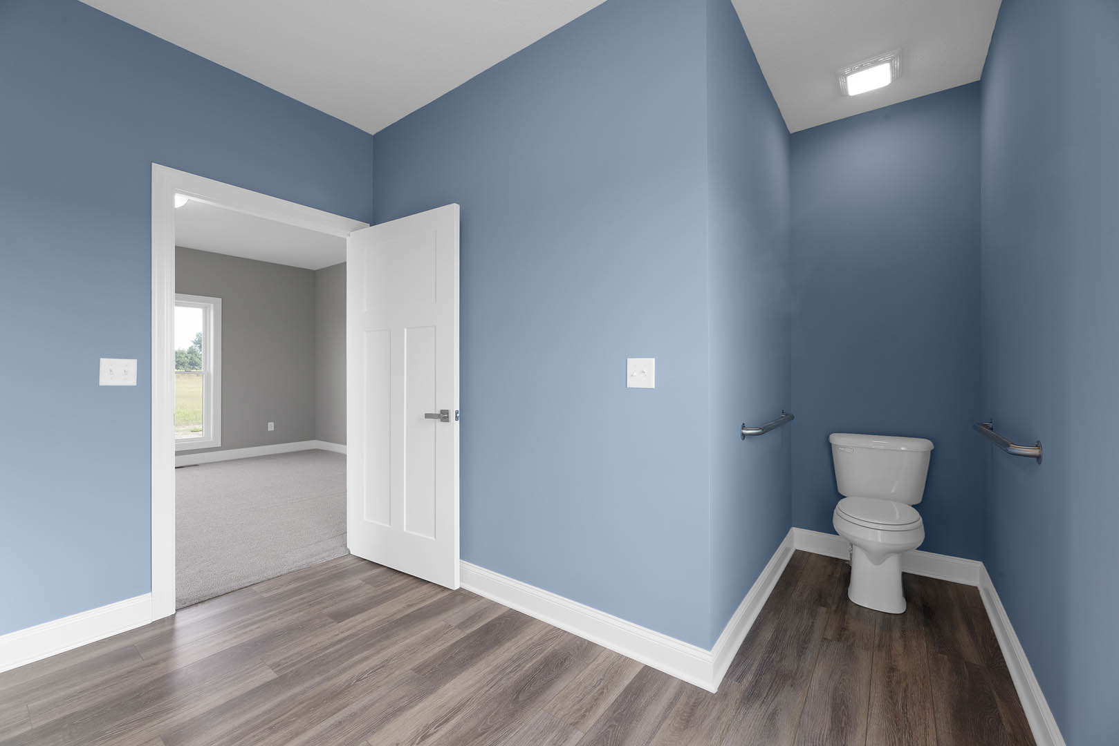 Modern bathroom with white toilet, wall-mounted metal handrail, tiled flooring, plaster walls, and window overlooking trees.