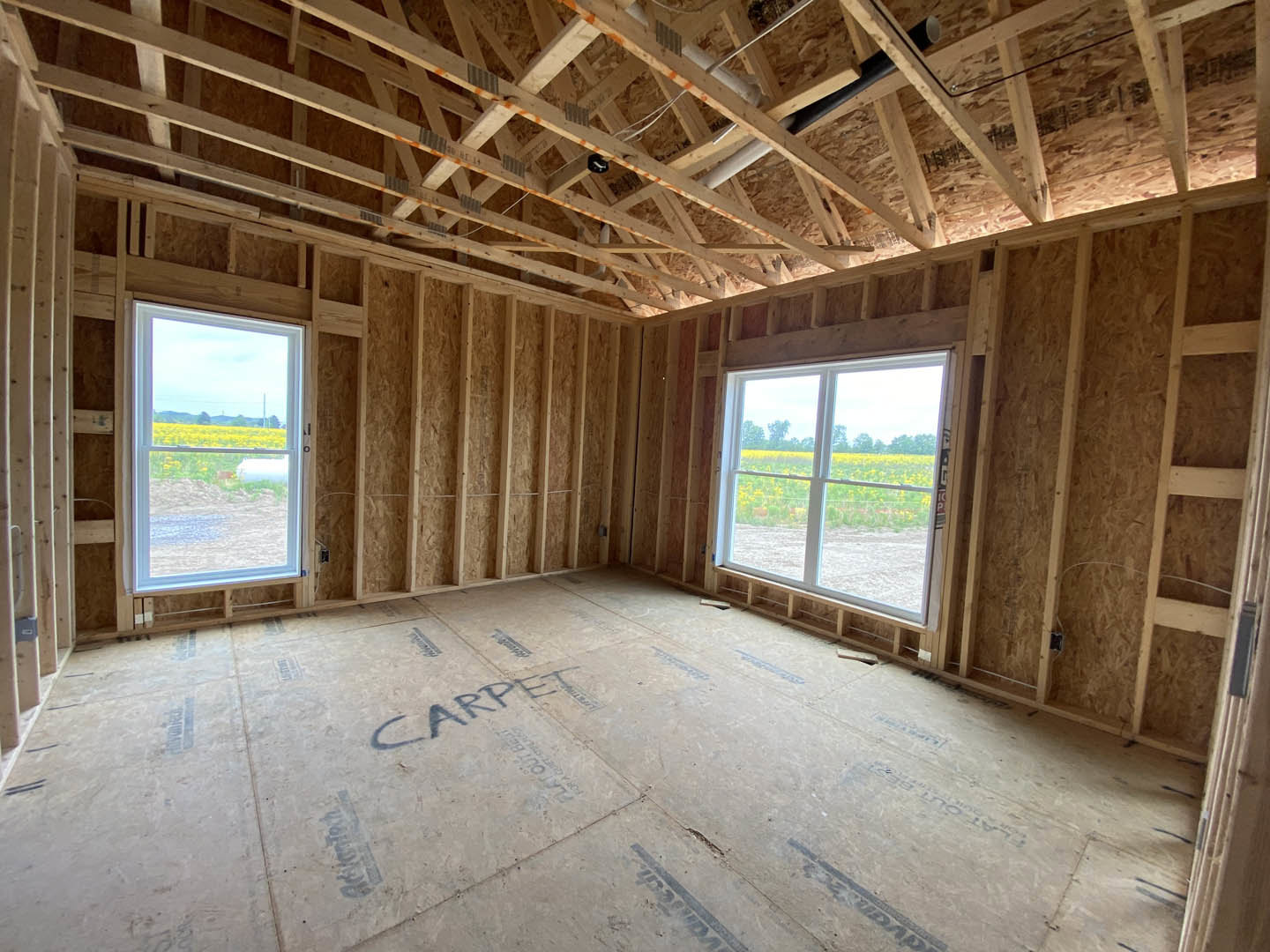 Spacious room featuring exposed wood ceiling beams, large windows overlooking a field of flowers, light-colored plank flooring, and visible ceiling pipes.
