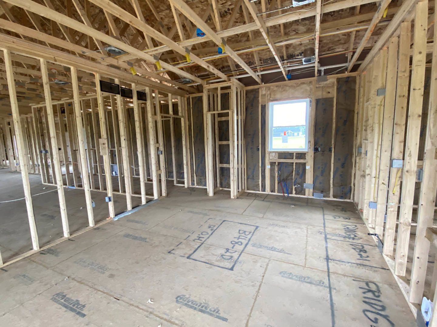 Room under construction with exposed wood framing, visible ceiling beams, concrete floor, and a window showing a blue house outside