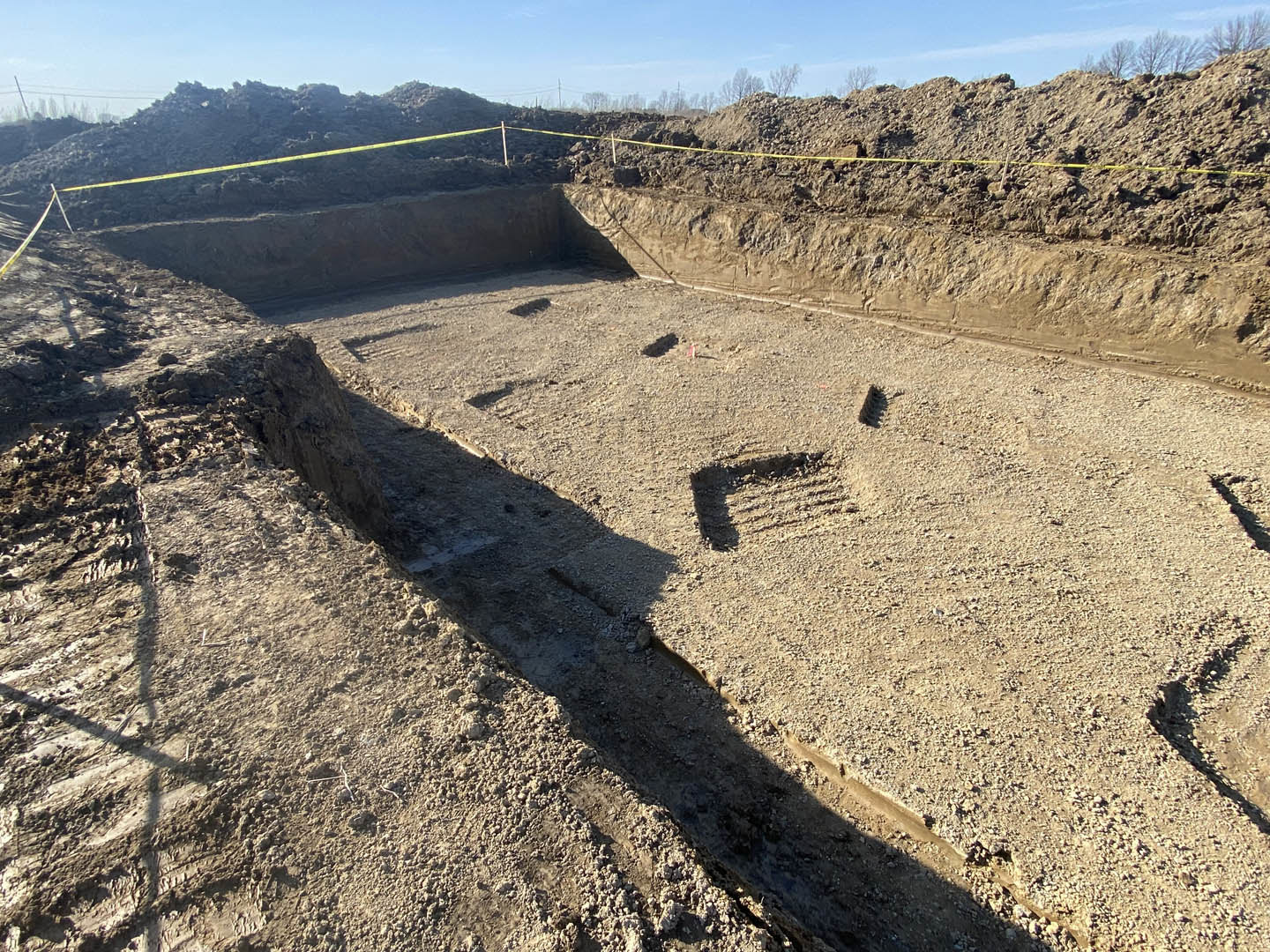 Dirt excavation pit surrounded by yellow caution tape, under blue sky with scattered clouds, tree in background, close-up of soil with visible footprint