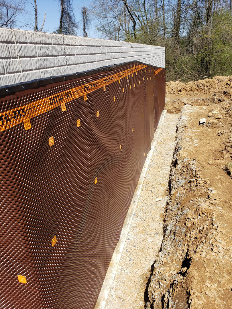Brown textured exterior wall with black mesh and yellow dots, adjacent to leafless trees and a white brick wall featuring black and brown stripes.
