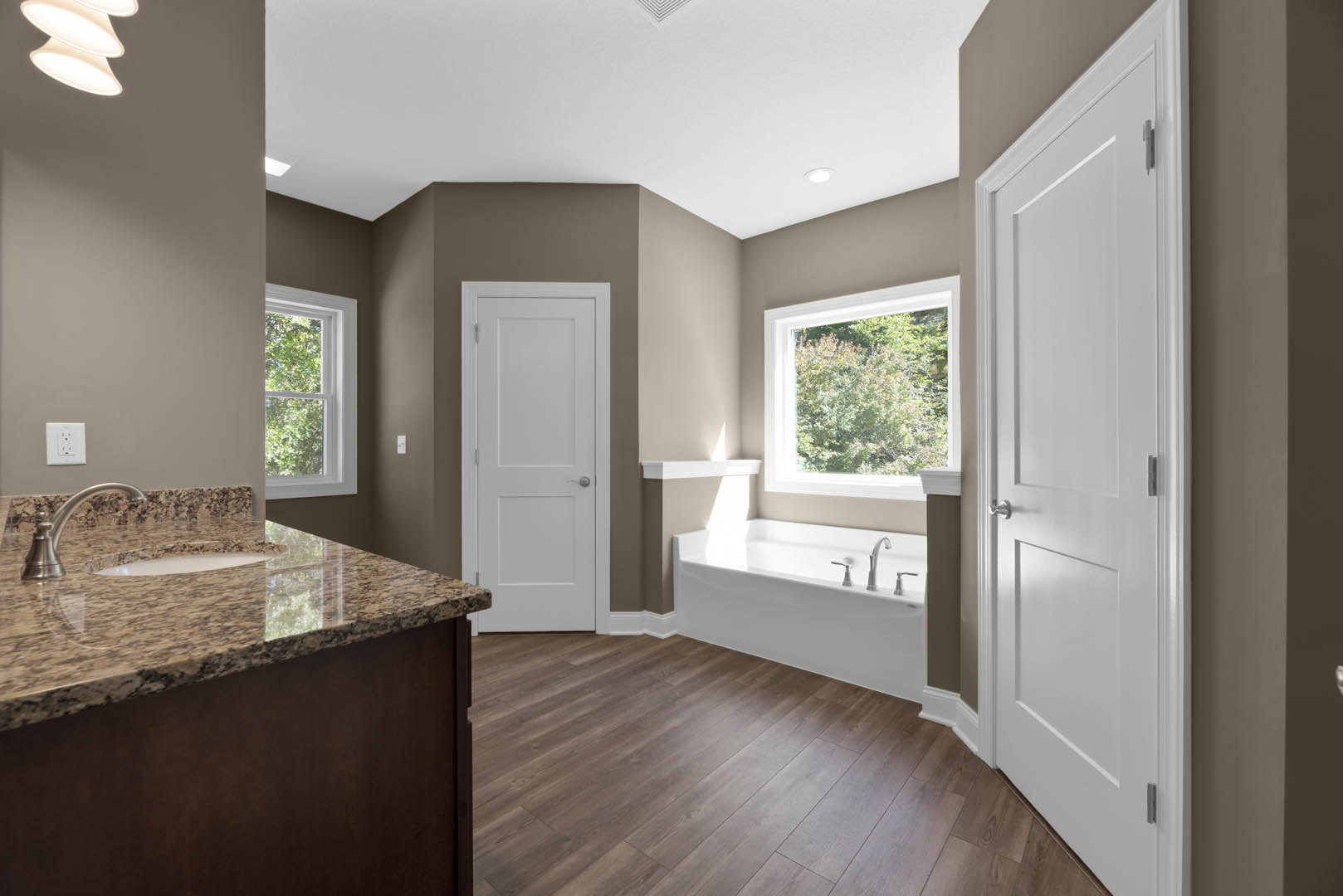 Bathroom featuring a marble countertop vanity, freestanding bathtub, white door with silver handle, window overlooking trees, white wall outlet, and recessed ceiling light