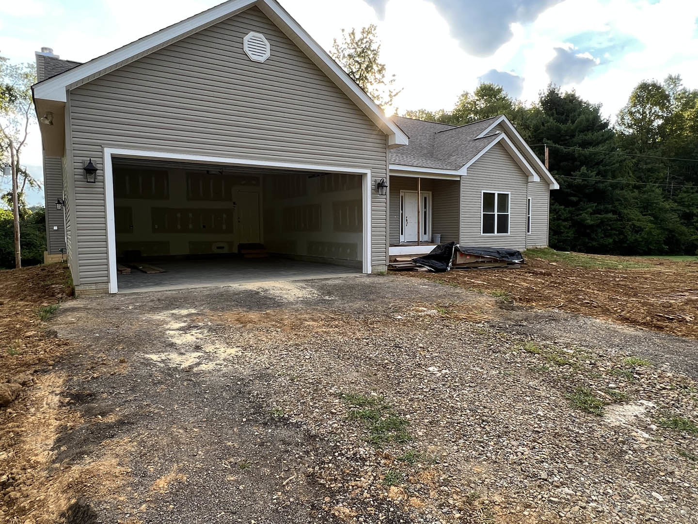 Grey exterior home with attached garage featuring a white paneled door, gravel driveway, white-framed window, and white vent on the facade; surrounded by trees and plants under a