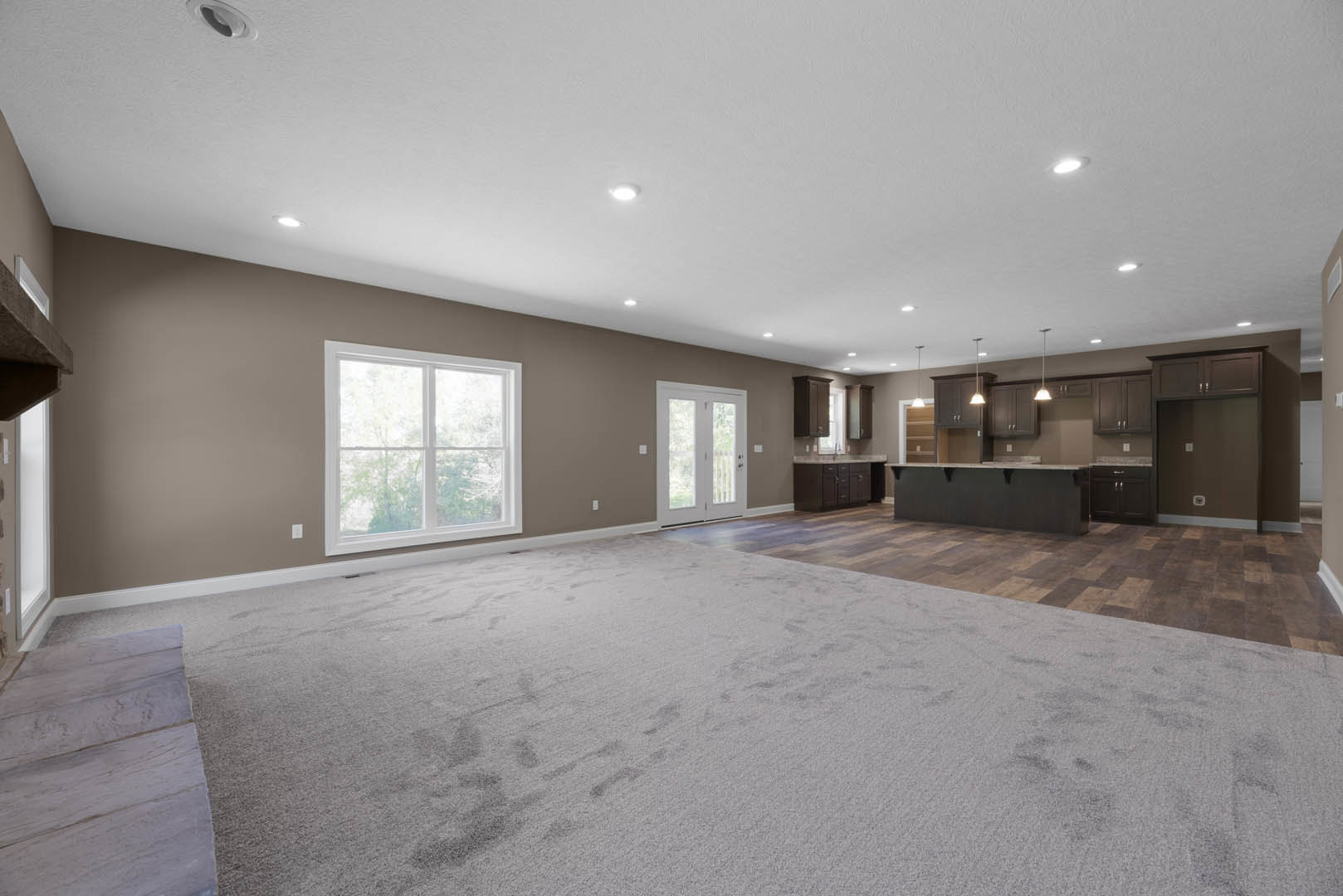 Open-concept kitchen and dining area featuring a large wooden table, white-framed window, glass-paneled white door, neutral carpet, recessed ceiling lights, and a black accent wall