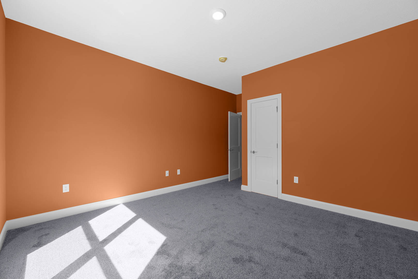 Empty room with orange painted walls, grey carpet flooring, white door with silver handle, ceiling light fixture, and a white carpet featuring a cross pattern.