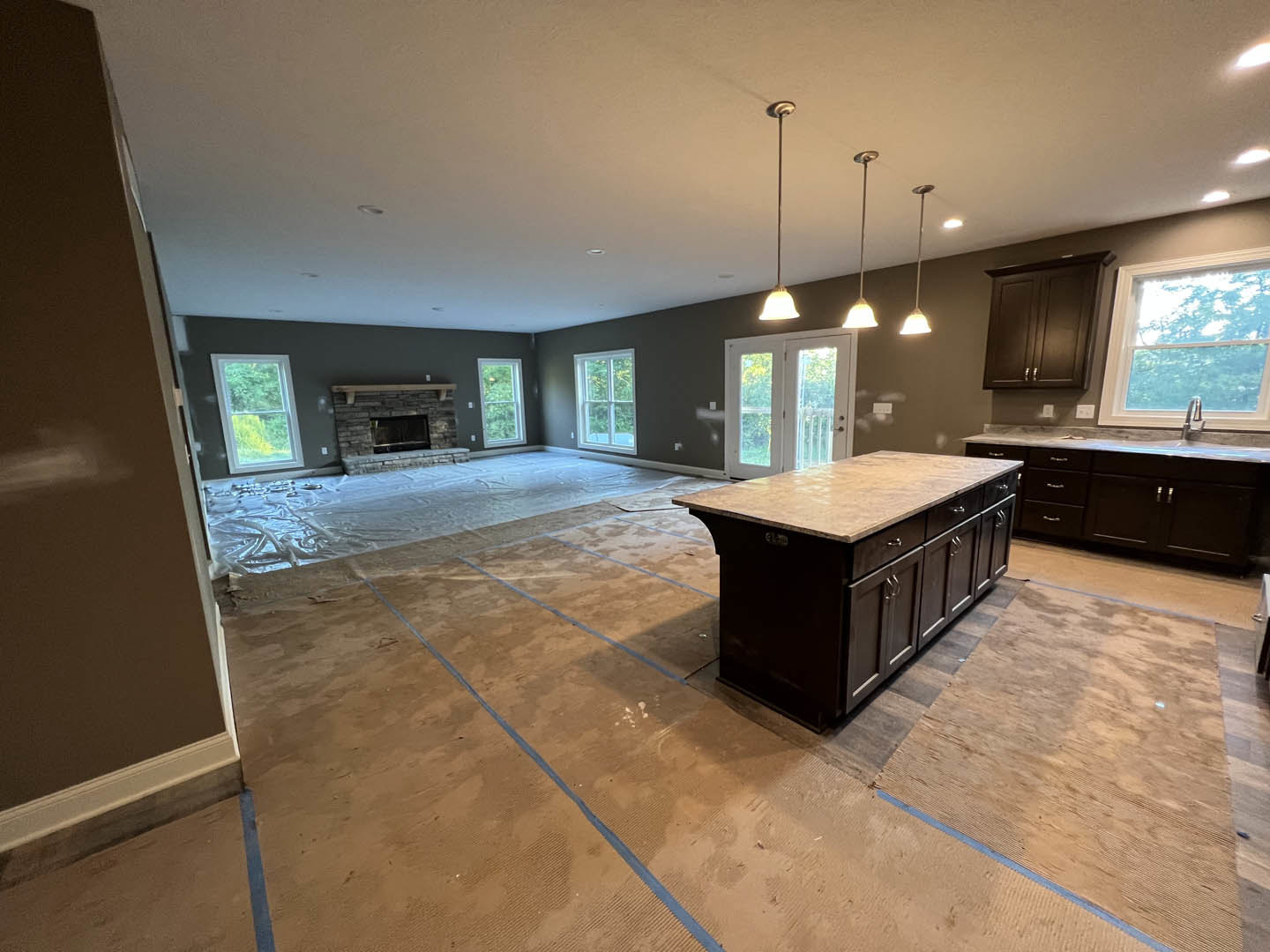 Marble-topped kitchen island, brick fireplace centered between glass-paneled double doors and white-framed windows, stainless steel sink beneath window, wood cabinetry and light
