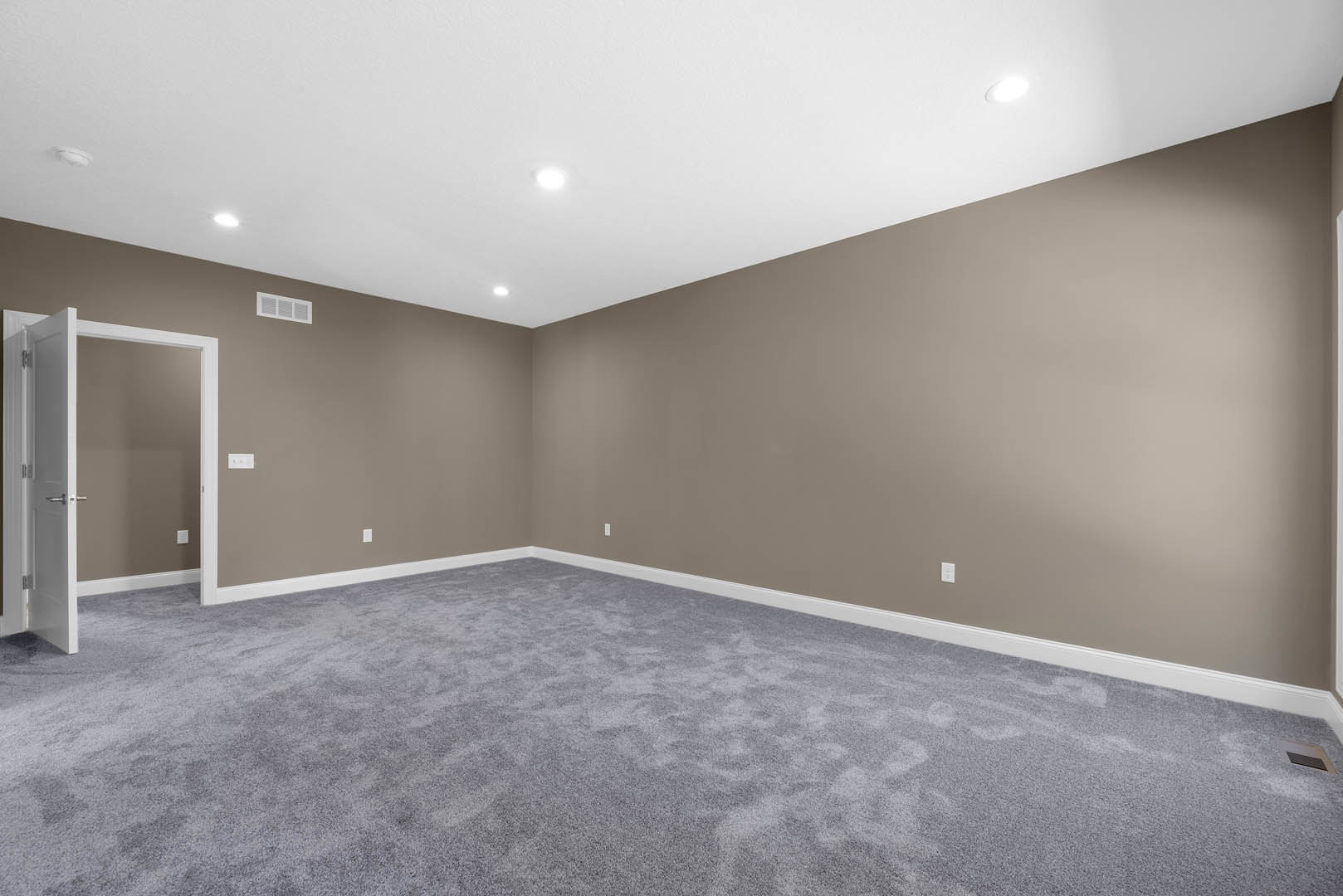 Grey carpeted floor, white ceiling with recessed lights, white walls, closed white door featuring a silver handle, empty interior room