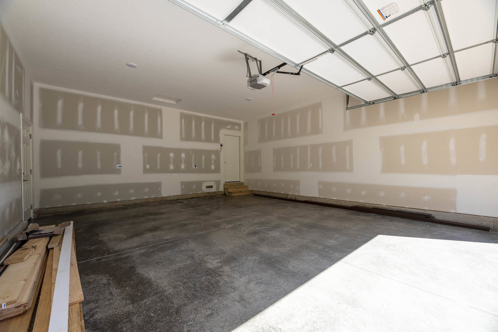 Garage interior with white painted plaster wall, white door featuring black handle, concrete floor illuminated by daylight, stacked wood planks near wall, ceiling with recessed