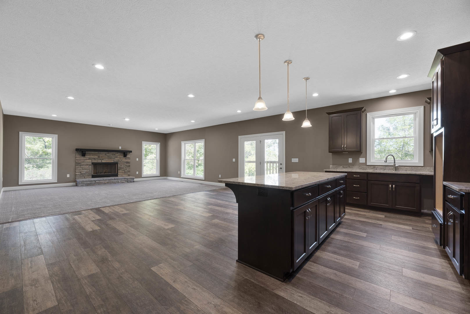 Spacious kitchen featuring a marble-topped island, stone fireplace, white cabinetry, wood flooring, and large windows overlooking trees