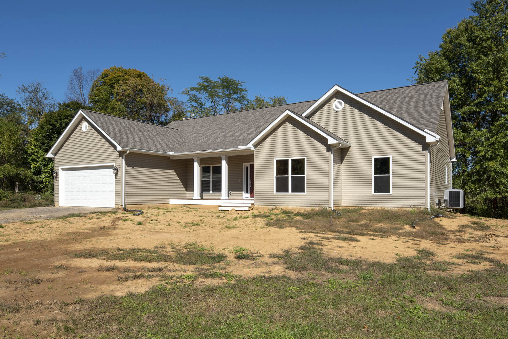 Two-story house with white siding, red front door, covered porch, white-framed windows, attached white garage door, grassy yard, dirt patch near entrance, central air conditioning