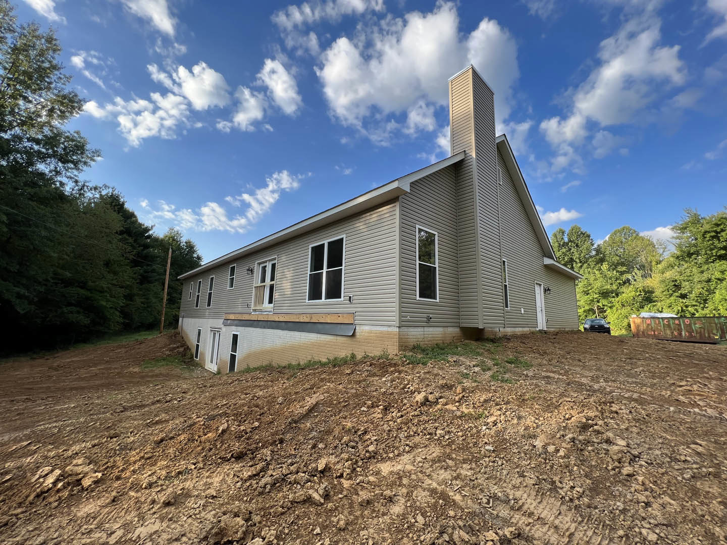Partially built house with exposed framing, white window frames, brick chimney, surrounded by dirt mound and mature trees under cloudy sky