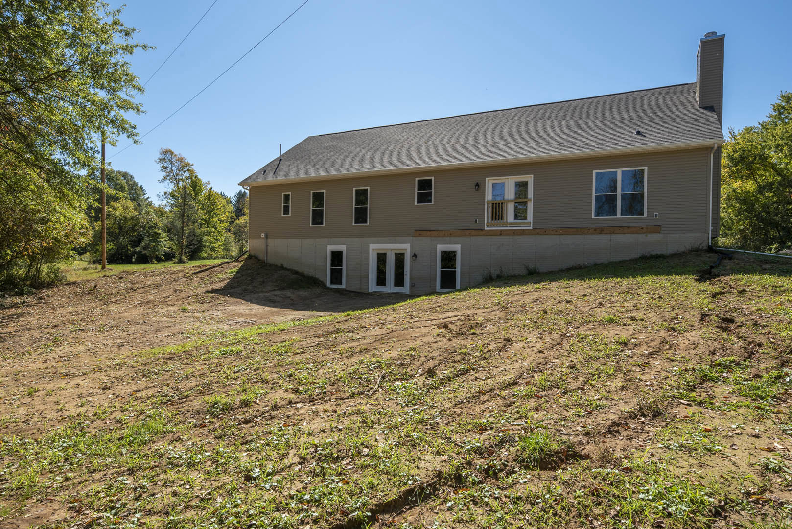 Two-story house with gray roof, white-framed windows, and glass double doors, set on a grassy lot with young plants and a hill rising in the background.