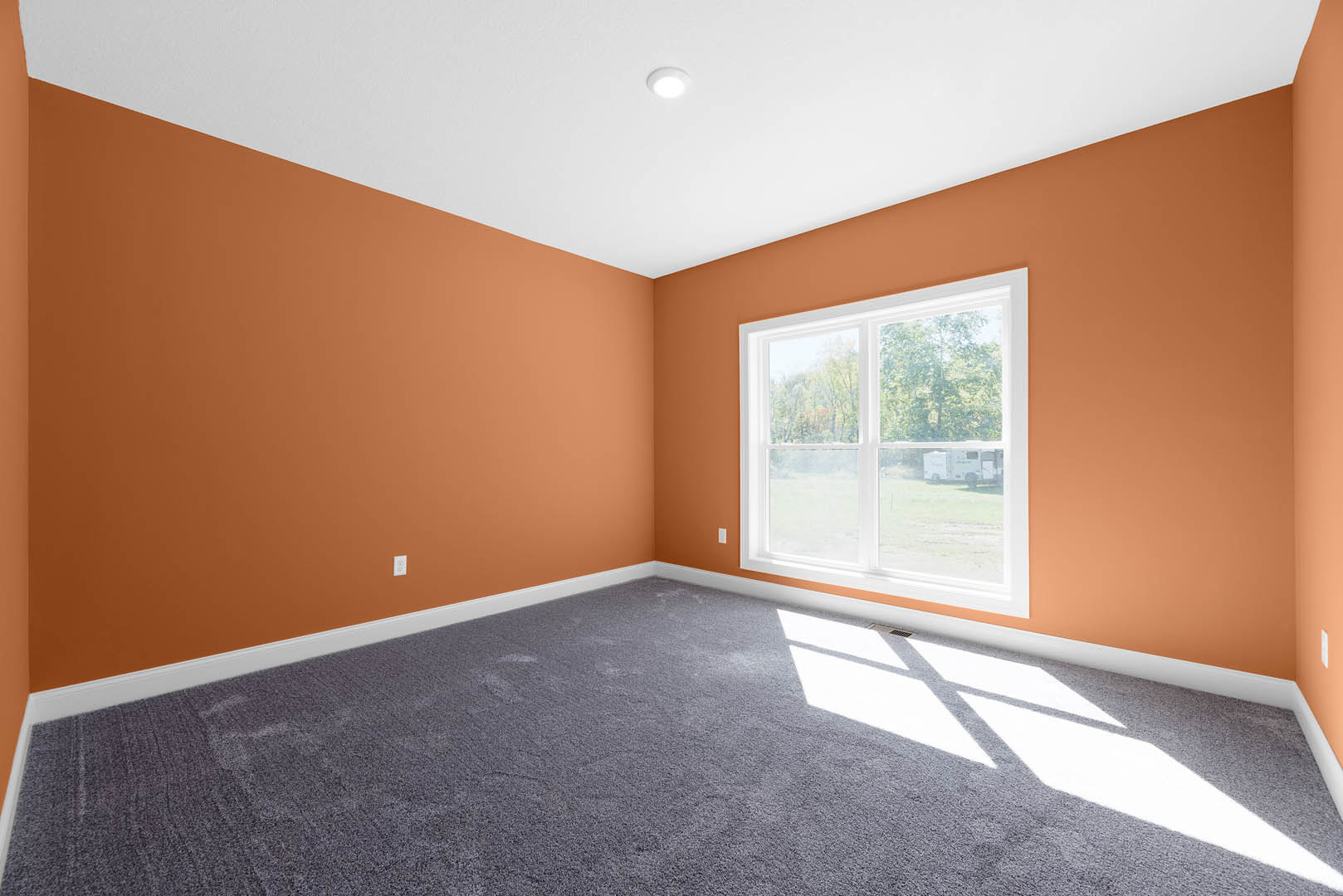 Empty room with orange walls, beige carpet flooring, white ceiling with recessed light, single window overlooking a camper outside