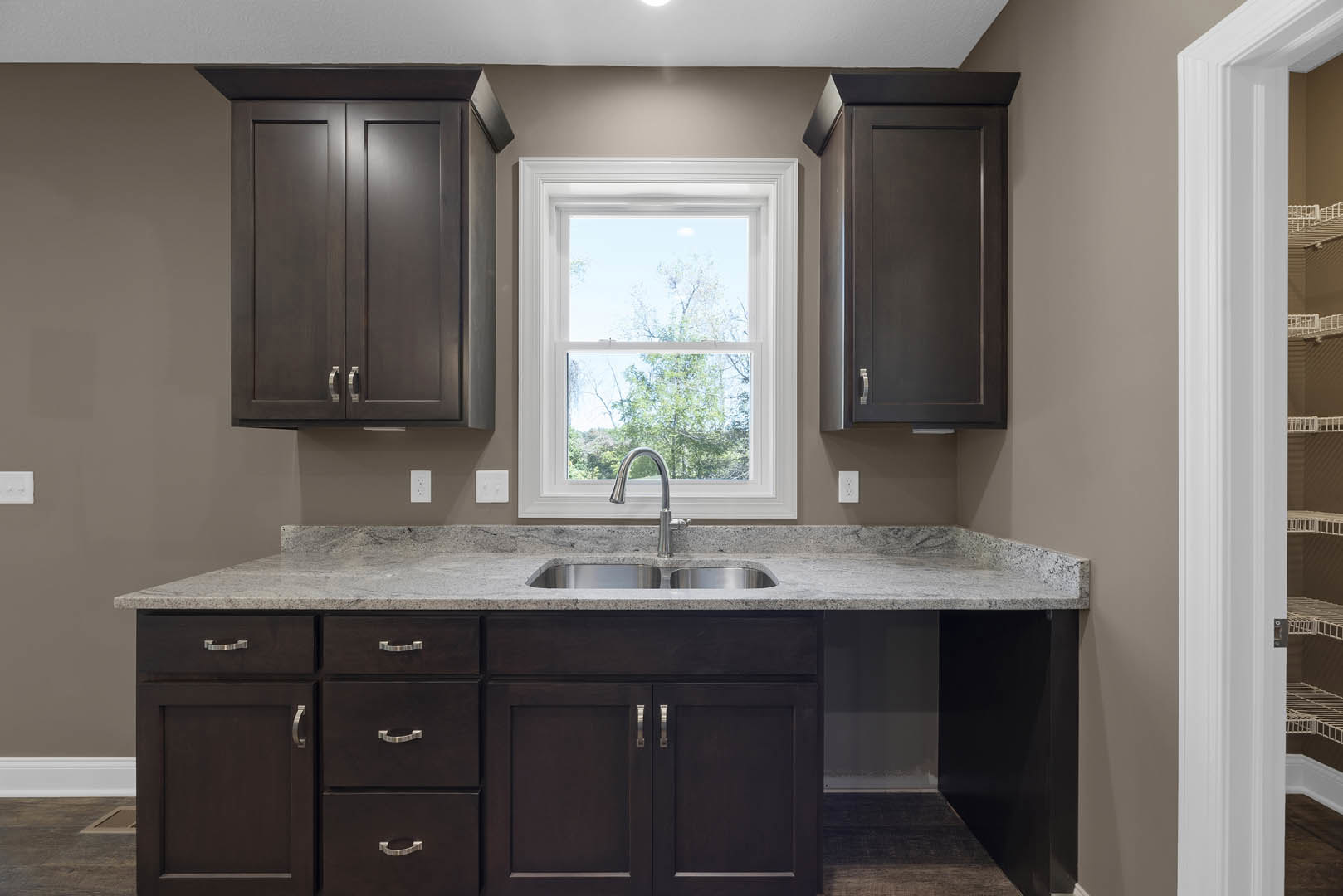 Dark wood kitchen cabinets, stainless steel sink beneath a window with trees visible outside, white countertops, black door with white trim, person standing beside a table