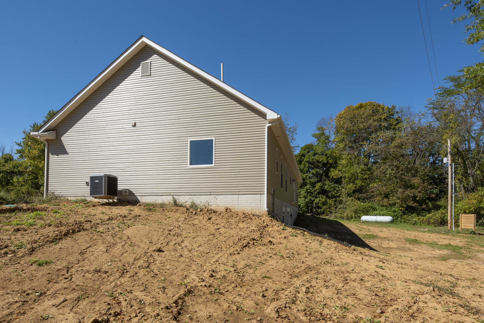 Two-story home with light siding, large windows, and black air conditioning unit, set behind a wooden fence on a grassy lot with a dirt hill and mature trees.