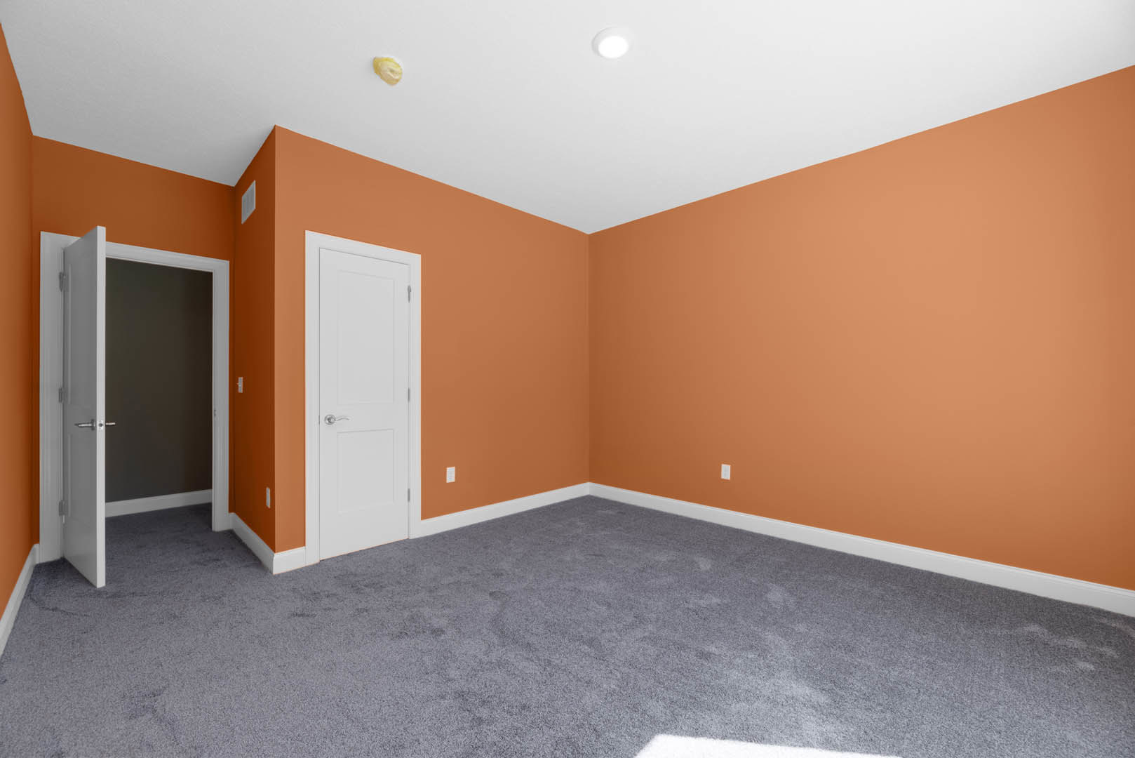 Empty room with orange painted walls, white door with silver handle, grey carpet flooring, and white ceiling