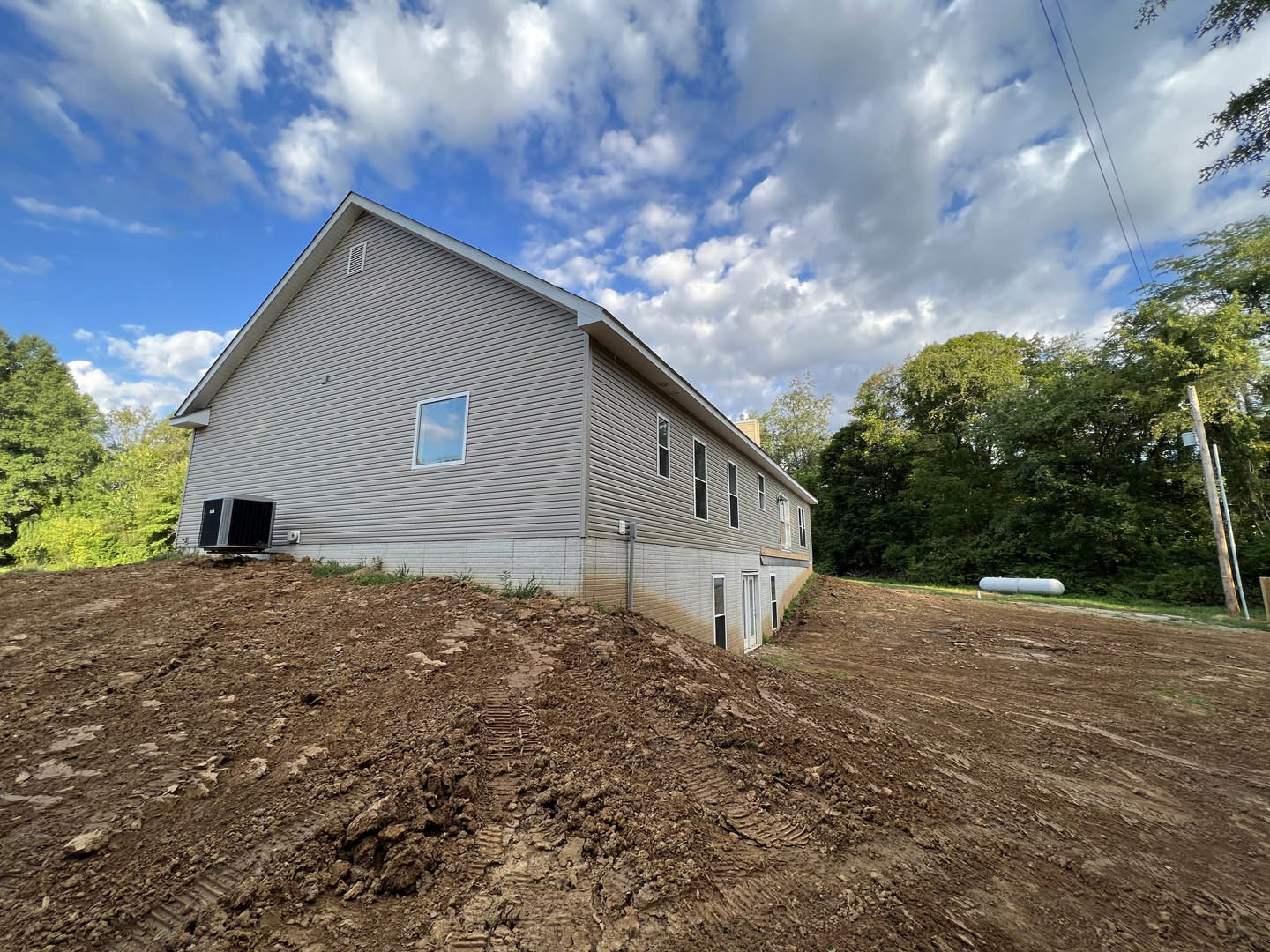 Modern house with light siding and large windows, situated on a dirt hill with visible tire tracks, surrounded by sparse grass and a rural landscape under a partly cloudy blue sky