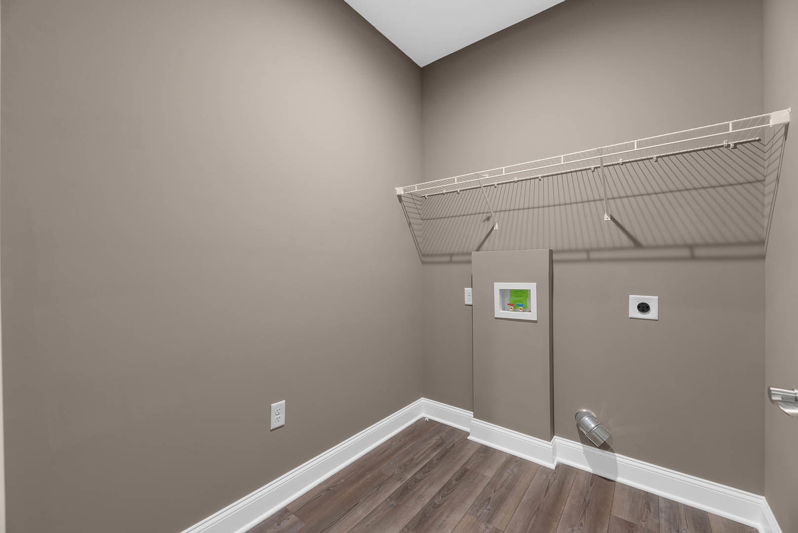 Wood floor with white baseboard, white shelving unit featuring a metal bar, white electrical outlet, and framed artwork with green and red paper against plaster walls
