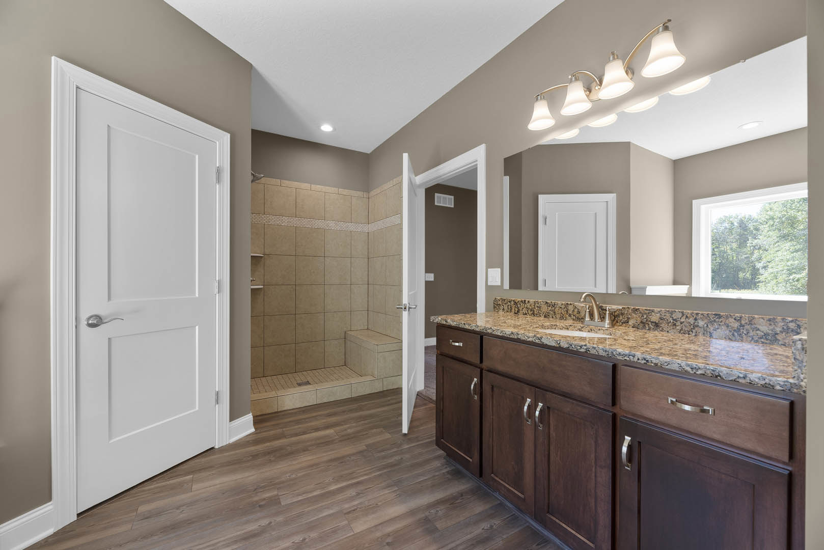 Bathroom featuring a marble countertop with undermount sink, tile-walled shower, white door with silver handle, row of wall-mounted lights, window overlooking trees, and light