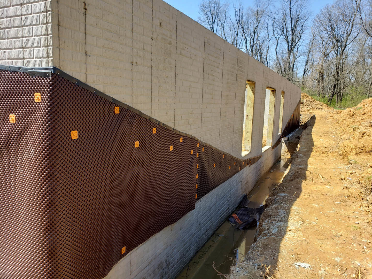 White exterior wall with rectangular windows, brown mesh panel, and circular hole near the ground; trees and blue sky visible in background.