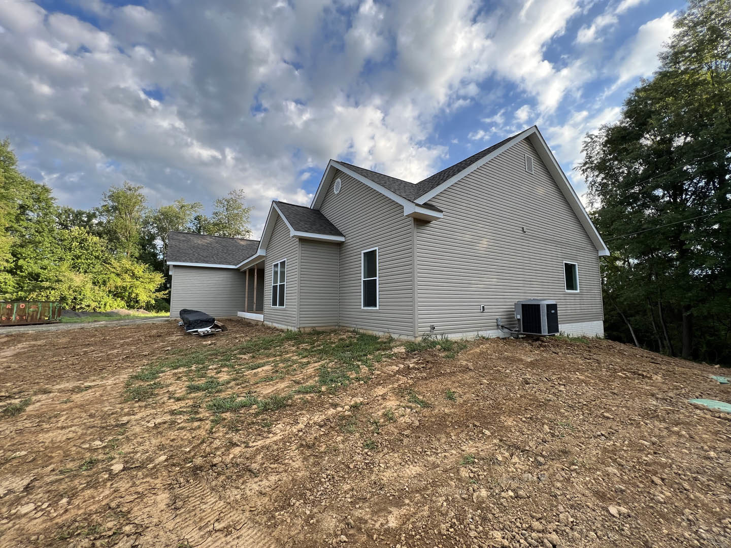 Two-story home with light siding, gabled roof, and large windows, surrounded by trees and a dirt hill in the foreground under a partly cloudy blue sky