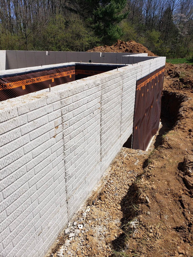 Red brick exterior wall with exposed foundation hole in dirt, surrounded by piles of soil, brown mesh, and nearby trees.