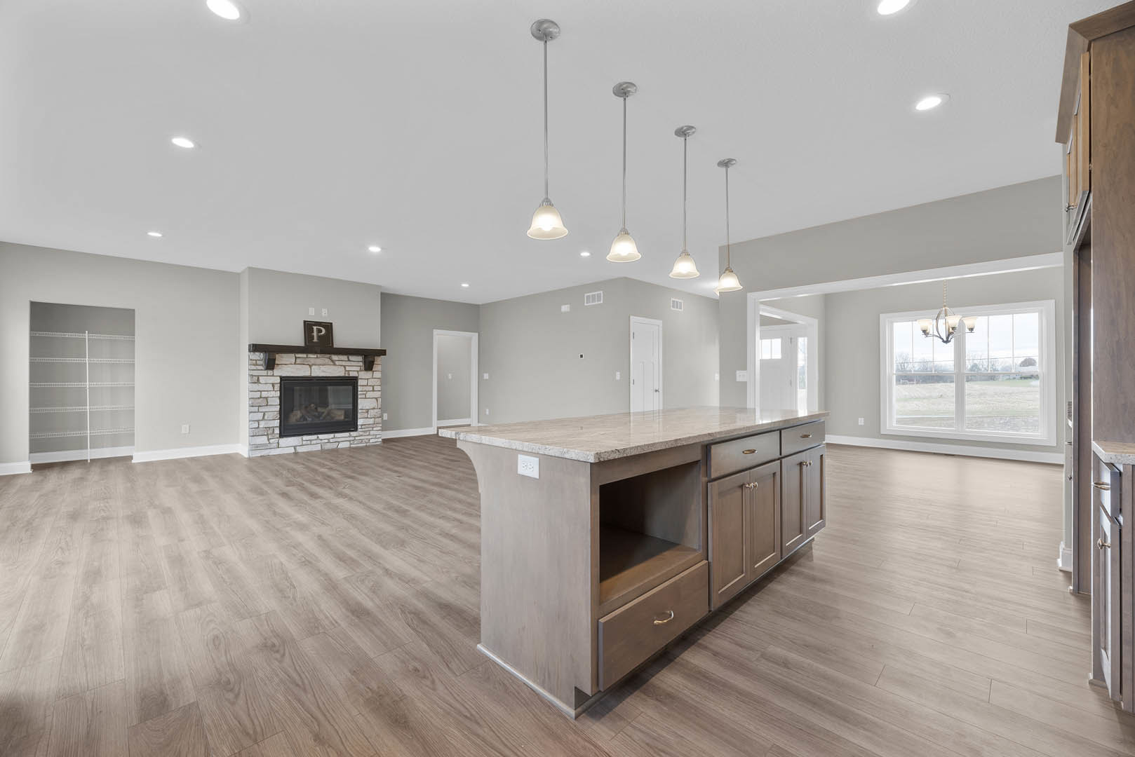 Spacious kitchen featuring a central marble-topped island with drawers, stone fireplace with glass window, white shelving with matching handles, large window with glass panel, and