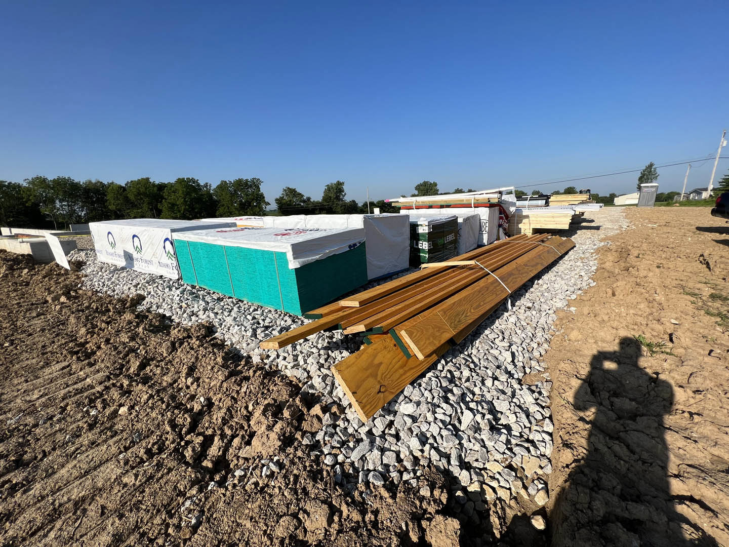 Stacked wood planks and building materials on rocky ground, blue rectangular object with white plastic covering, clear blue sky, surrounding trees, shadow of person holding camera